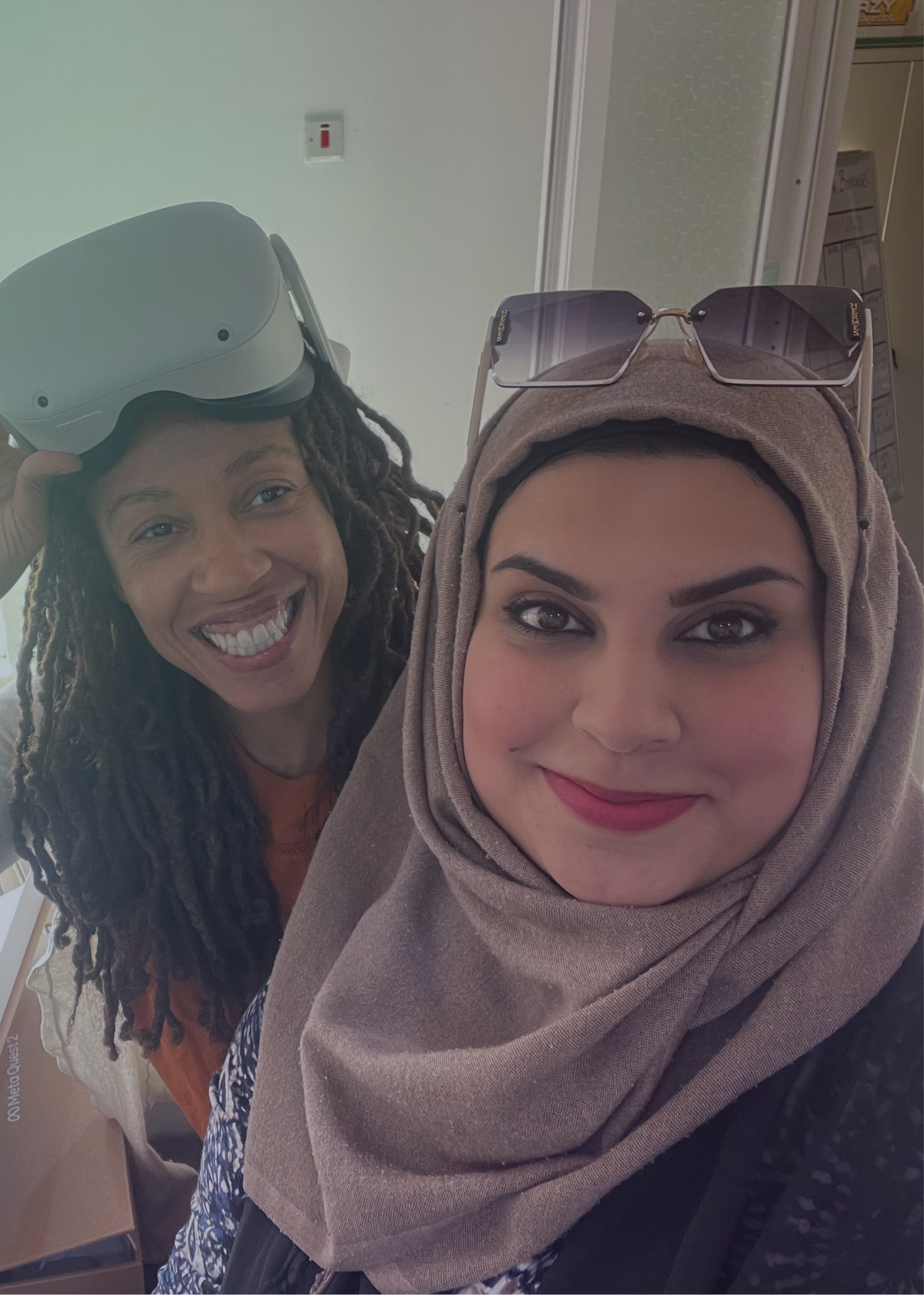 Two women smile in a selfie. One wears VR goggles on her head.